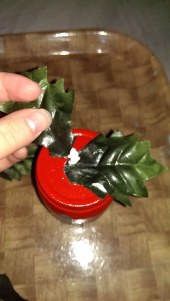 Adding plastic holly leaves to red baby food jar.